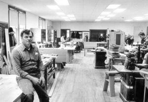 A man sits on a workbench in a spacious and very clean woodworking shop as others work at stations nearby, amid tools and bright ceiling lights.