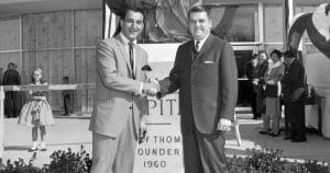 Two men in suits shake hands and smile by a building and statue, with formally dressed people behind. 1960s black-and-white photo.