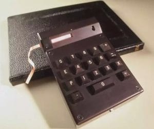 A vintage black calculator with large push buttons and a paper tape sits on top of a closed black leather notebook on a light surface.