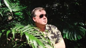 A man in sunglasses and a tropical shirt stands among lush plants, gazing sideways as sunlight casts shadows on his face.
