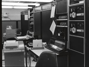 A black-and-white photo of a vintage computer room with large PDP computers, tape reels, control panels, an old typewriter, a rotary phone, and a single desk chair in the foreground.
