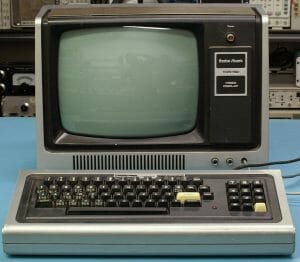 A vintage Radio Shack TRS-80 computer with a built-in monitor and separate keyboard sits on a blue table near electronic gear.