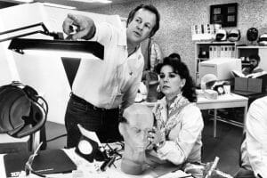 A man points as a woman examines a mannequin head in a busy office or lab, with gear, helmets, and people working behind them.