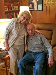 An older woman stands and smiles, embracing an older man who sits in an armchair. Both look happy in a warm, wood-paneled room filled with books, a lamp, and family photos on the wall.