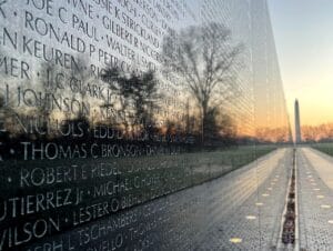Close-up of the Vietnam Veterans Memorial wall with engraved names, reflecting trees and the Washington Monument in the background at sunrise, creating a serene and respectful atmosphere.