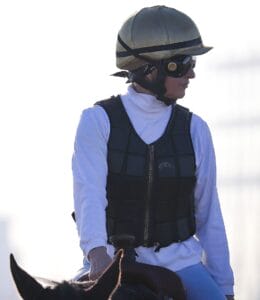 A woman in a protective vest, white turtleneck, tan helmet, and goggles sits on a horse, facing sideways against a bright backdrop.