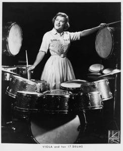 A woman in a vintage dress smiles behind an elaborate drum set with many drums, holding drumsticks; caption: “Viola and her 17 drums.”.