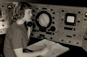A woman at a vintage control panel with rotary dial and screens holds a phone to her ear, writing in a large open notebook.