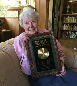 An elderly woman with white hair sits on a couch holding a USGS award plaque reading Virginia Norwood in a cozy room with bookshelves.