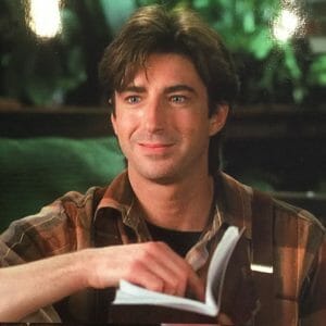 A smiling man with brown hair in a plaid shirt flips through a small book, with blurred greenery visible in the background.