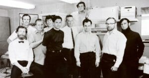 Eleven men pose in a computer lab with vintage equipment in the background. Black-and-white photo.