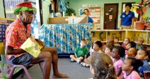 An older man in a festive hat and colorful shirt reads to kids sitting on the floor in a classroom as two adults watch from the background.