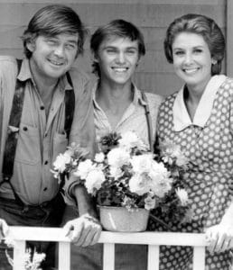 Three actors stand smiling on a porch behind a white railing, with a potted bouquet of flowers in front of them. The two men wear casual shirts; the woman wears a patterned dress and has short hair.