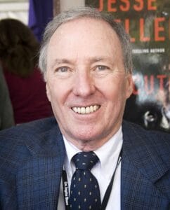 A smiling older man with thinning gray hair in a blue checkered suit, white shirt, and navy polka-dot tie, seated indoors.