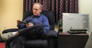 A man in glasses and a blue shirt sits in an armchair, examining a circuit board next to a vintage CUBIS CEC computer.