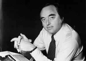 A man in a dress shirt and polka dot tie sits at a desk with his hands folded, looking at the camera. A keyboard and documents are visible in front of him. The photo is in black and white.