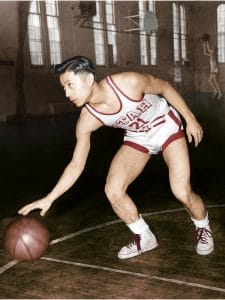 A basketball player in a vintage uniform, labeled STATE 15, dribbles a basketball on an indoor court. He has short dark hair and wears white high-top sneakers. Two other players are visible in the background.