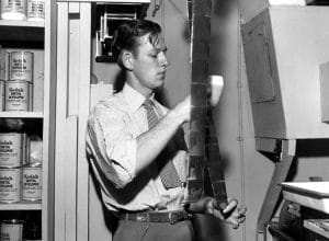 A man in a striped shirt and tie examines a strip of photographic film in a darkroom, surrounded by cans of Kodak developer and equipment.