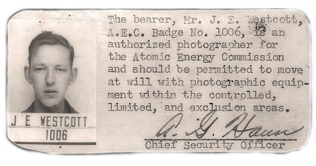 A black-and-white ID badge showing a young man’s photo and stating J.E. Westcott, badge number 1006, as an authorized photographer for the Atomic Energy Commission, signed by the Chief Security Officer.