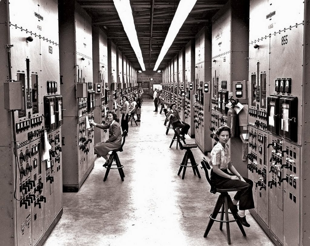 Black and white photo of women sitting on stools, operating large control panels in a long, industrial room lined with equipment on both sides. Overhead lights run down the center of the ceiling.