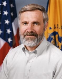 A middle-aged man with short, light brown hair and a full gray beard wears a light-colored shirt, posing in front of the American and U.S. Public Health Service flags.