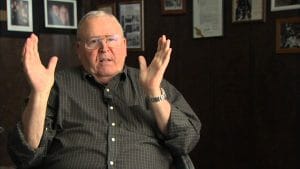 An older man in glasses and a button-up shirt sits indoors, gesturing as he talks. Framed photos and documents are on the dark wall behind him.