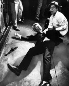 A man in a suit lies on the ground with his arm outstretched, another kneels by him, concerned. Several people stand nearby.