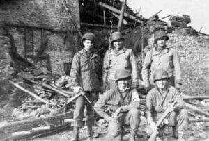 Five soldiers in military uniforms and helmets pose for a photo in front of a damaged, partially collapsed building with scattered debris. Two soldiers are kneeling, while three stand behind them, all holding equipment or rifles. It is in black and white, from World War II.