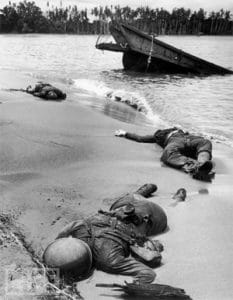 Three fallen soldiers lie on a sandy shore near a partially sunken boat in shallow water, with trees visible in the background across the river.