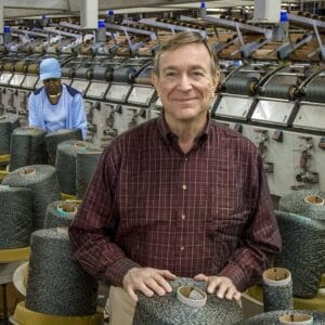 A man in a plaid shirt stands smiling in a factory, with large spools of thread in front of him. In the background, a worker in a blue uniform operates machinery.