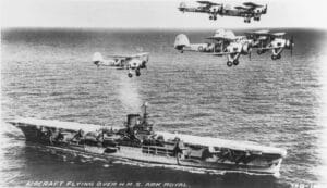 Six vintage biplanes fly in formation above the British aircraft carrier HMS Ark Royal as it sails on the ocean, in a black-and-white historical photograph.
