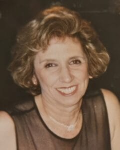 A smiling woman with short, wavy, light brown hair wears a sleeveless, sheer black top and a delicate necklace, posing against a dark background.