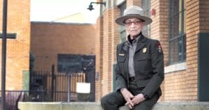 A park ranger in uniform and hat sits on a low concrete wall in front of a brick building, looking toward the camera with a slight smile.