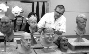 A man works on a sculpted head in a workshop, surrounded by various detailed busts and masks of different characters displayed on stands. Shelves with more masks are visible in the background.