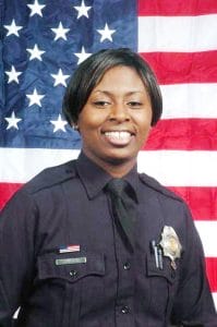 A smiling police officer in uniform stands in front of a large American flag backdrop.