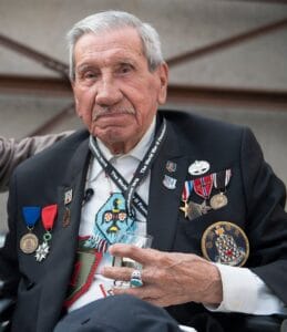 An elderly man with white hair and a mustache wears a suit jacket adorned with various medals and pins. He holds a small glass and looks at the camera with a calm expression.