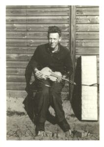 A man sits outdoors in front of a wooden wall, holding a violin. He is dressed in dark clothing and appears to be looking at the camera. A rectangular object is positioned beside him on the ground.