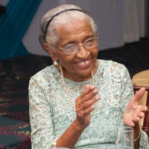 An elderly woman with gray hair, glasses, and pearl jewelry smiles and claps her hands. She is wearing a light green lace dress and is seated indoors at an event.