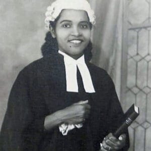 A woman dressed in a judge’s robe and white collar, wearing a white judicial wig, smiles while holding a book in one hand, standing in front of a patterned backdrop.