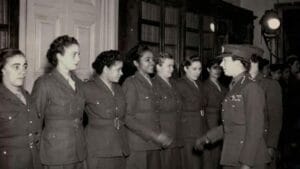 A group of women in WWII military uniforms stand in a row, facing a female officer who appears to be inspecting or greeting them. The setting is indoors, with shelves of books in the background.
