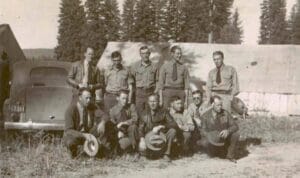 A group of men in uniform pose outdoors in front of canvas tents and a 1930s vintage car. Some are standing, some kneeling, all holding hats. Tall pine trees are visible in the background on a sunny day.