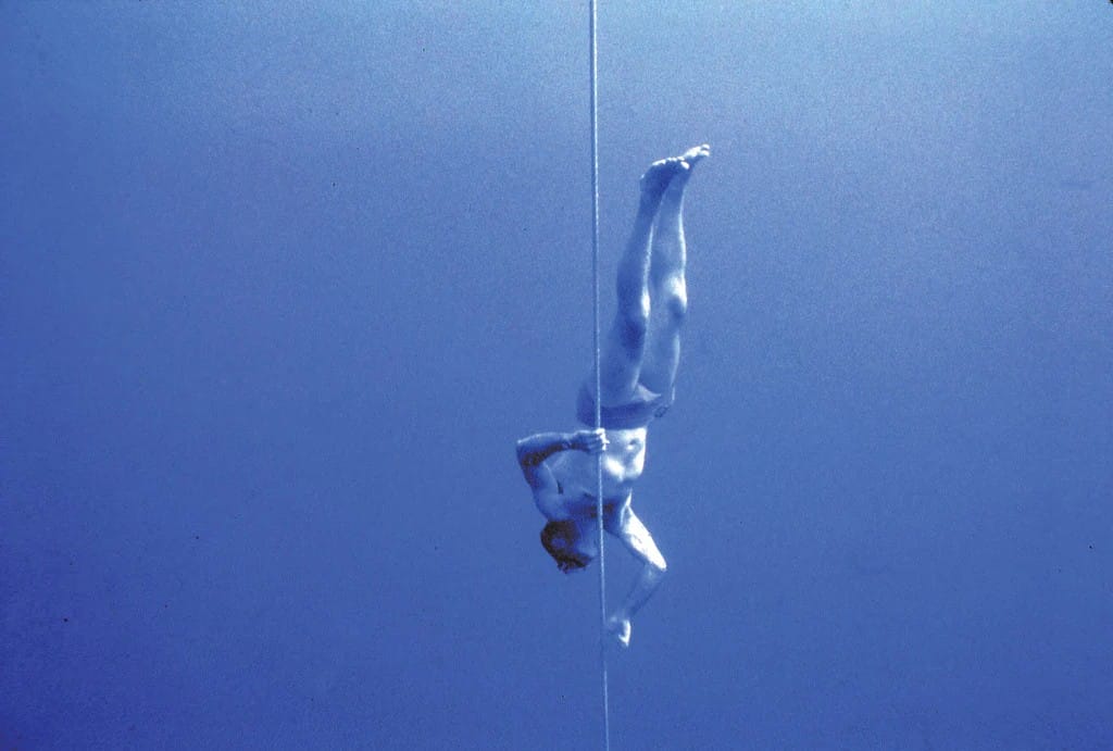 A person underwater, upside down, descending along a vertical rope in deep blue water, with legs extended upward and one hand holding the rope.