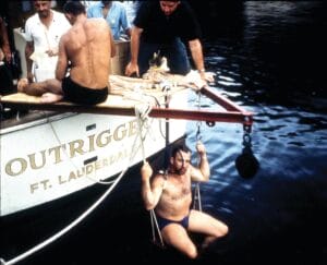 A man in swim trunks sits on a swing-like seat hanging off the back of a boat named Outrigger FT. Lauderdale. Two other men are on the boat, and the water is dark below them.