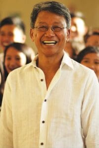 A smiling older man wearing glasses and a white button-up shirt stands in front of a blurred group of people, creating a warm and joyful atmosphere.