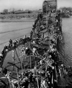 Crowds of people climb over the damaged, twisted metal structure of a partially collapsed bridge spanning a river, with more people gathered on the riverbanks in the background, under a clear sky.