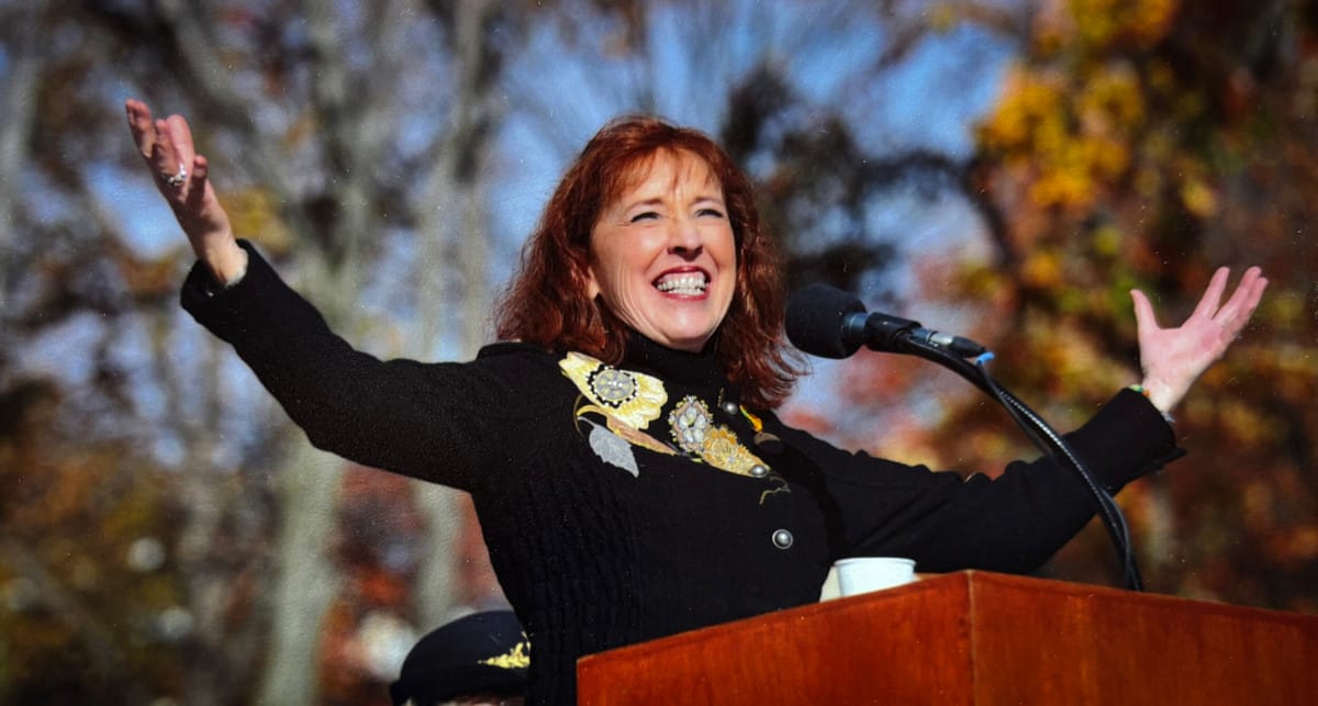 A woman with red hair stands at a lectern outdoors, smiling broadly with arms raised. She wears a black coat decorated with floral embroidery. Autumn trees with orange leaves are visible in the background.