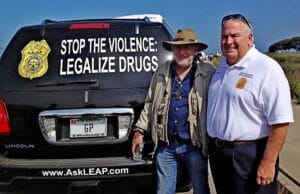Two men stand smiling beside a black SUV with a sign reading STOP THE VIOLENCE: LEGALIZE DRUGS and a LEAP badge logo on the rear window. The man on the right wears a white polo shirt, the other a hat and glasses.