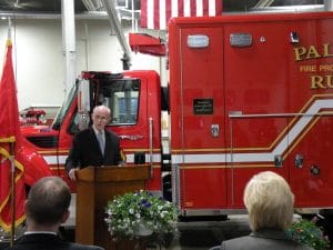 An older man speaks at a lectern in front of a red emergency vehicle inside a fire station, with an American flag overhead and an audience seated in the foreground.