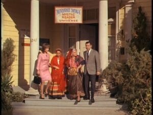 Four people, two women in modern clothes, a woman dressed as a fortune teller, and a man in a suit, walk out of a building with a Madame Mona Mystic Reader Universe sign above the entrance.