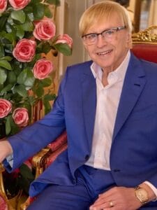 A smiling man with blond hair and glasses, wearing a blue suit and white shirt, sits on a red and gold ornate chair next to a bouquet of pink roses.